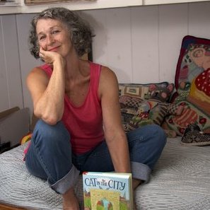 Jill Weber sits cross legged in blue jeans and a pink tank top holding the book "Cat in the City"