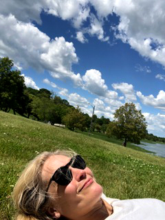 Image of author Lenaye Marsten laying in a green field on a sunny day with a few fluffy clouds in a deep blue sky.