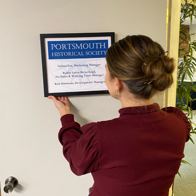 Museum employee in a burgundy shirt faces awat from the camera as they hang up a blue and white office sign on a door.