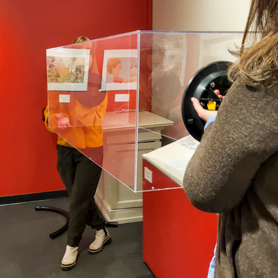 Two museum professionals remove a rectangular plexiglass bonnet from a display case using suction cup grips.