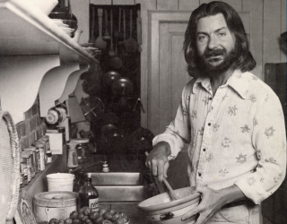 Image of Buddy Haller, holding a ceramic bowl and stirring something with a wooden spoon. Buddy is wearing a button-up shirt with a floral patter. Buddy stands in front of a stovetop with shelving above it. 