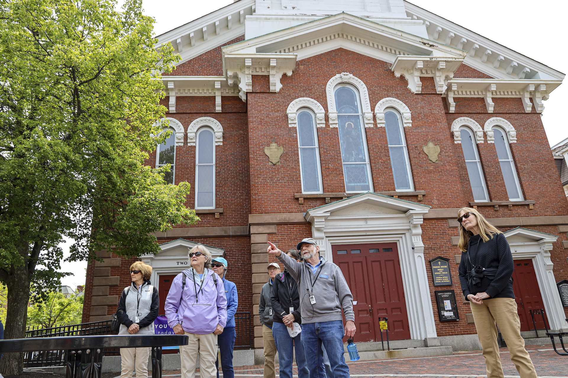 Walking tour of nine people standing in Market Square with an old fashinoed lamppost next to them and a tall brick church with a white steeple in the background.