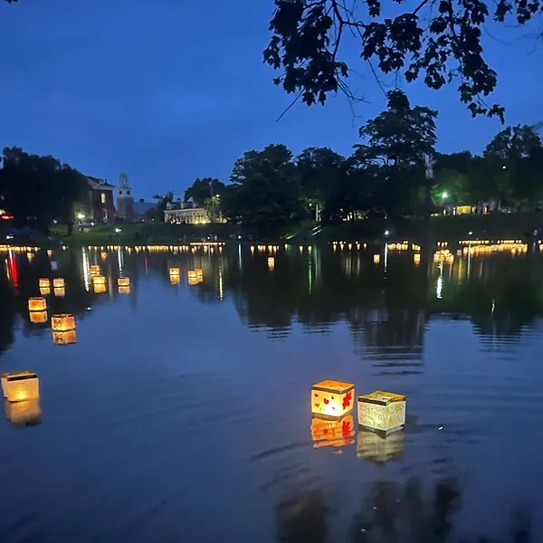 Two paper lanterns sit on a large pond of water as dusk settles in. Trees line the far side of the pond.