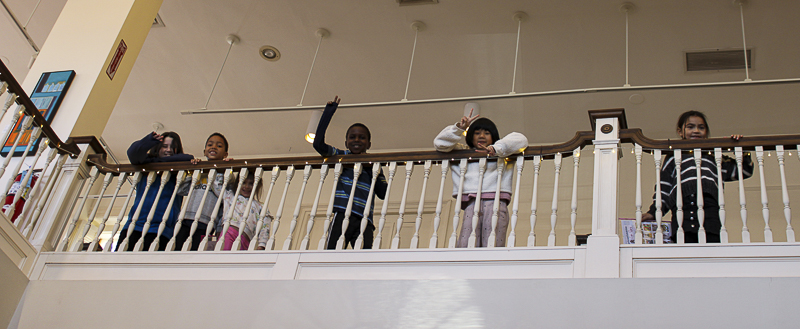 Five children wave down at the photographer from the upstairs gallery over a white wooden balustrade. 