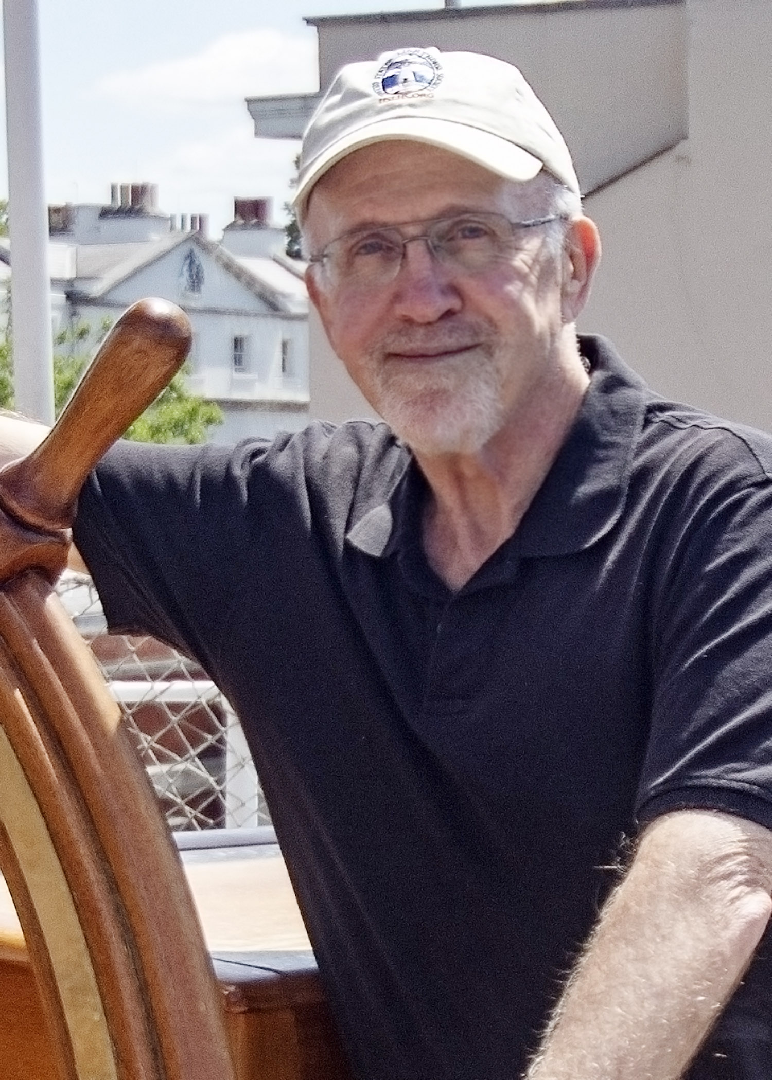 Author Jeremy D’Entremont. A white man sits behind a wooden captain's wheel with glasses and a white baseball cap on. 